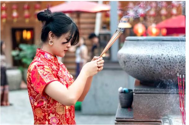زنی با لباس قرمز جشن چینی، چوب های بخور را با سر خمیده در دست دارد و آنها را به خدایی تقدیم می کند.